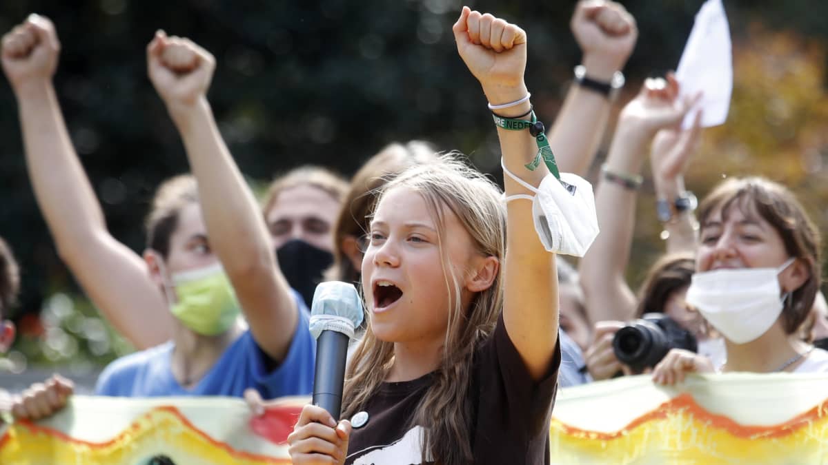 Greta Thunberg håller upp en arm i luften medan hon håller tal på en klimatdemonstration. I bakgrunden demonstranter i munskydd.