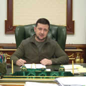 Volodymyr Zelenskyj sitter vid ett skrivbord. Han tittar framåt. I bakgrunden Ukrainas flagga.