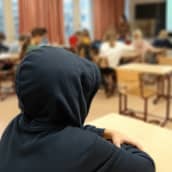 En elev sitter med huvan uppdragen på sin mörkblå munkjacka, långt borta från den övriga klassen.