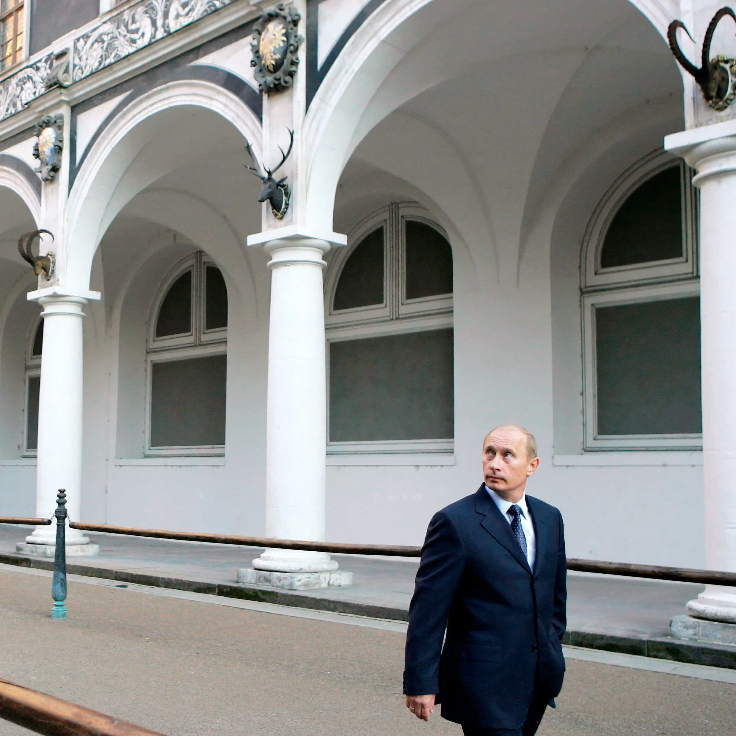 Vladimir Putin kävelee tummansinisessä puvussa ja kravatissa pylväskäytävämn edustalla ja katselee ylös kuvan vasempaan reunaan.