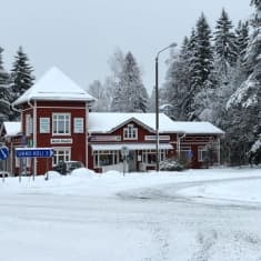 Lunta maassa, taustalla punainen talo, lumisia puita, liikennemerkkejä. 