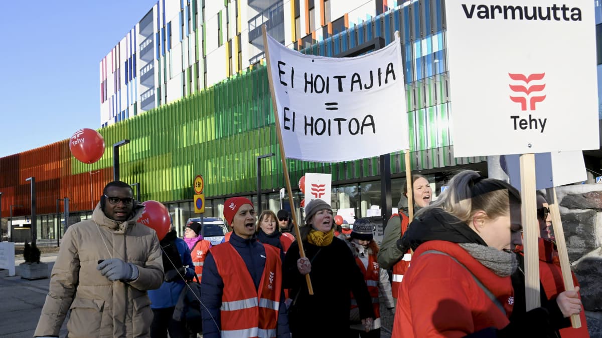Ett demonstrationståg utanför en byggnad med färgglad fasad. Människorna i tåget har röda västar och röda ballonger.