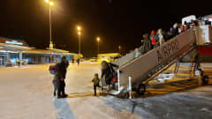 Ihmisiä poistumassa lentokoneesta portaita pitkin lumisessa maisemassa. 