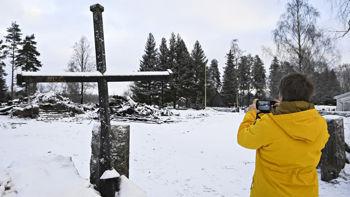 Keltaiseen talvitakkiin pukeutunut nainen kuvaa Rautjärven kirkon raunioita ja tulipalon jälkeen tapaninpäivänä portin pieleen nostettua kirkon ristiä 28. joulukuuta 2022. 