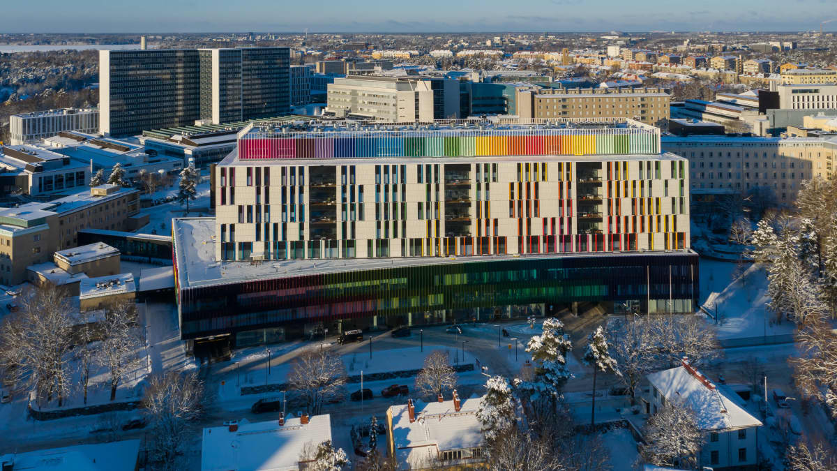 Uusi lastensairaala Helsingissä ilmasta kuvattuna. On talvi, maassa on lunta ja aurinko paistaa kirkkaasti. Lastensairaalan seinän yläosassa näkyy paljon eri värejä. 