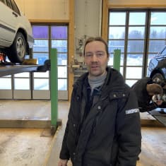 Katsastusmies seisoo hallissa. Hallissa on kaksi autoa, joista toisen renkaita tutkii toinen katsastusmies.