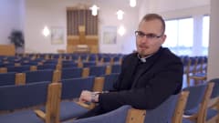 En präst sitter på en stol i en stor sal och pratar.