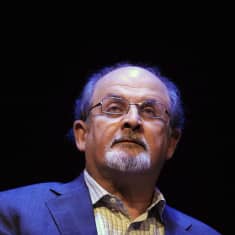 Salman Rushdie seisoo valokeilassa tummaa taustaa vasten.