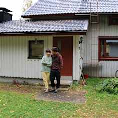 Colegeasuun pukeutuneet tummahiuksiset mies ja nainen seisovat vanhan, valkoisen rintamamiestalon edessä ja nojaavat toisiinsa.