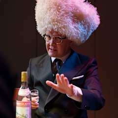 En man i kostym och enorm skinnmössa dricker vodka.