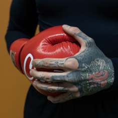 Miehen vasemmassa kädessä on punainen nyrkkeilyhanska ja oikeassa, paljaassa kädessä on tatuointeja, joissa näkyy punaiset nyrkkeilyhanskat.