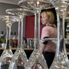 Rivissä seisoo väärinpäin olevia kirkkaita cocktail-laseja. Taustalla näkyy epätarkasti Misa Latva-aho.