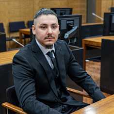 A young man with short dark hair, mustache and goatee sits in court, wearing a dark suit.