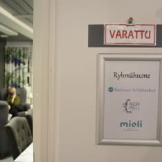 Ovessa näkyy NuoriMieli-keskuksen ryhmähuoneen kyltti, ja taka-alalla kaksi ihmistä istuu ryhmähuoneessa keskustelemassa.
