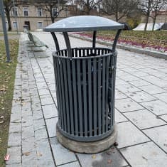 Tummanharmaa metallinen roska-astia kivetyllä jalkakäytävällä.