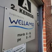 Harmaassa ovessa kyltti, jossa on työterveys Wellamon logo sekä aukioloajat.