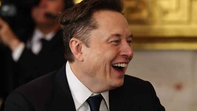 En man i mörk kostym och slips skrattar i sidoprofil.