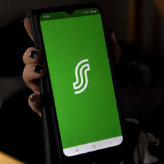 Käsi pitää matkapuhelinta, jonka ruudulla S-Pankin logo vihreällä pohjalla.