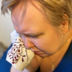vaalean, lyhytttukaisen naisen eli ääneellä itkijä Marjo Akkasen kasvot kuvattuna yläviistosta. Nainen painaa poskelleen vaaleaa, kirjailtua itkuliinaa.