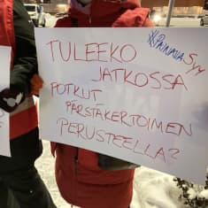 Valkoinen kyltti, jossa lukee "#painava syy, tuleeko jatkossa potkut pärstäkertoimen perusteella?". Kylttiä pitelee punatakkinen ihminen, ja maa on luminen.
