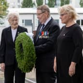 Alla Hoskonen, Kyösti Tomperi ja Mari Ekmark seisovat muistomerkin edessä. Kyöstillä seppele kädessä.