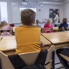 Lektion pågår i klassrum. I förgrunden två elever, ena i gul t-shirt och andra i blårandig t-shirt. Ryggsäckar på stolsryggarna.