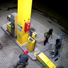 Valvontakamerakuva ylhäältäpäin, kolme kasvonsa peittänyttä varasta on kaatanut tankkausaseman automaatin ja seisovat sen ympärillä.