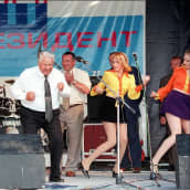 Boris Jeltsin tekee tanssieleitä lavalla kravatti kaulassa ilman puvuntakkia. Kuvan vasemmassa reunassa näkyy mieslaulaja punaisessa paidassa ja liiveissä. Kaksi naista minihameissa tanssii Jeltsinin oikealla puolella. Kankaalla taustalla lukee isoin kirjaimin: Meidän presidenttimme.