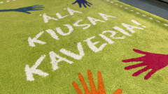Matto koulun käytävällä, matossa värikkäitä käsiä ja teksti: Älä kiusaa kaveria!