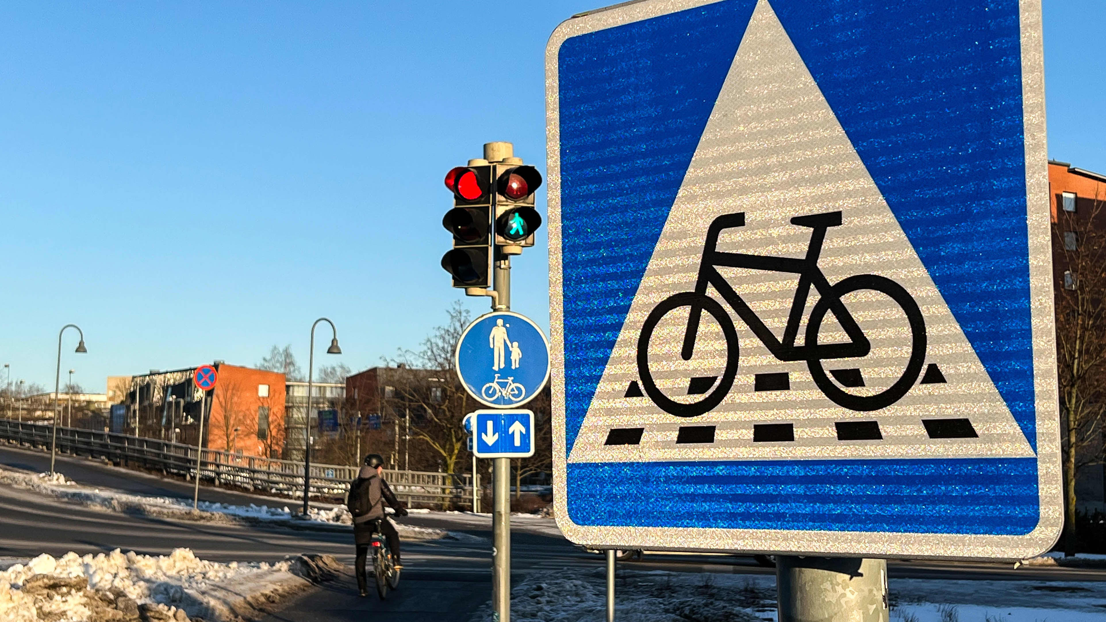 Polkupyöräilijä odottaa valojen vaihtumista Turun Aninkaistensillan kupeessa. Liikennemerkki B7, väistämisvelvollisuus pyöräilijän tienylityspaikassa. Neliönmallisessa sinipohjaisessa merkissa on valkoisen kolmion sisällä polkupyörän kuva sekä katkoviivoitus osoittamassa pyörätien jatketta.
