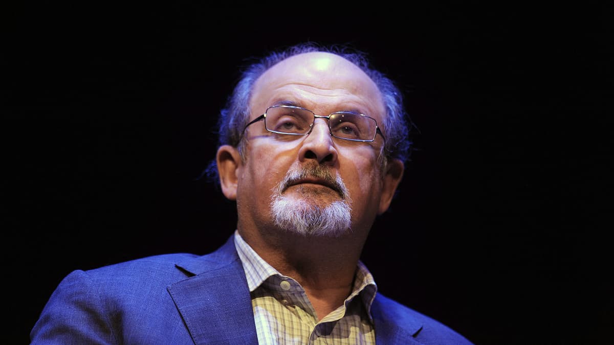 Salman Rushdie seisoo valokeilassa tummaa taustaa vasten.