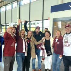 Kahdeksan ihmistä seisoo rivissä kädet kohotettuna. Oikeassa reunassa seisovalla miehellä on päällään Latvian signeerattu pelipaita.