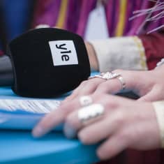 Kahden saamenpukuisen naisen kädet on laskettu pöydälle Ylen makuullaan olevan mikrofonin viereen.