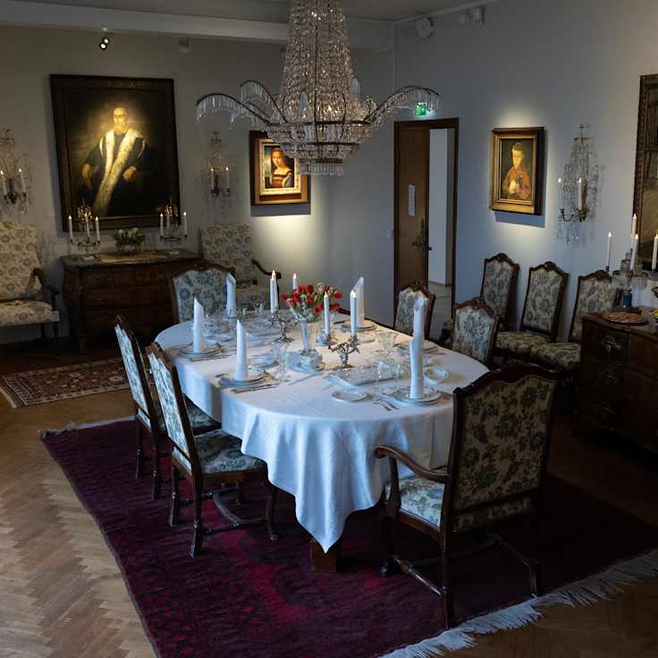 Tyylikäs ruokasali, jossa on elegantti kynttiläkruunu, kattauksena valkoinen pöytäliina ja koristeellisia tuoleja. Seinillä on suuria taideteoksia.