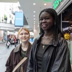 Vaaleahiuksinen lyhyt nuori nainen katsoo vierellään seisovaa pitkää tummaihoista nuorta naista hymyillen kauppakeskuksen aulassa.