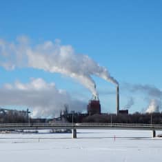 Iso tehdas, jonka piipuista tupruaa harmaata savua sinistä taivasta vasten. Tehtaan edessä silta, joka johtaa joen yli.