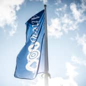 En blå flagga med Attendos logo vajar i en flaggstång. I bakgrunden syns blå himmel med ett lätt molntäcke.