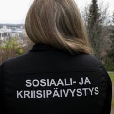 Nainen katselee syksyistä kaupunkia sosiaali- ja kriisipäivystys työntekjän takki yllään. 