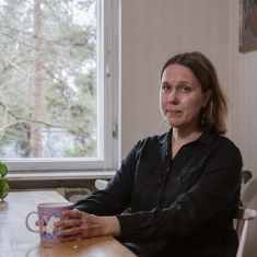 Helsinkiläinen Saana Hansen istuu ikkunan ääressä pöydän edessä ja katsoo kameraan käsissään violetti muumimuki.