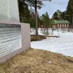 Vasemmalla muistomerkki, jossa on metallinen laatta ja siinä teksti: "Kemijoen uittoyhdistys 1901-1991". Taustalla näkyy ränsistynyt punainen rakennus.
