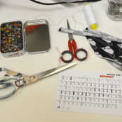 Pöydällä tekstiilikäsitöiden välineitä, esimerkiksi sakset, lankarulla, nuppineuloja.