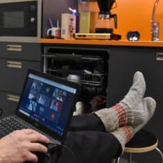 A man wearing woolen socks, feet up, holding a laptop, in a virtual meeting