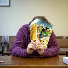 A person sitting at a table covering their face with an Aku Ankka magazine.