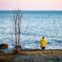 Nainen rannalla keltaisessa toppatakissa katselee kohti horisonttia.