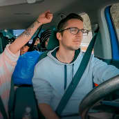 Kuva henkilöauton sisältä tuulilasista päin katsottuna. Kuskin paikalla isä Joachim Walhelm, etupenkillä äiti Patricia Walhelm. Äiti tuulettaa, on nostanut kätensä ilmaan riemuisasti. Takapenkki on täysi lapsista ja tavaroista.