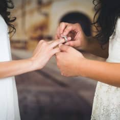 En kvinna i bröllopsklänning sätter en ring på fingret på en annan kvinna i bröllopsklänning.