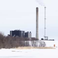 Lämpövoimalaitos talvimaisemassa rantapuiden ja kukkulan takana sekä kaksi isoa savupiippua.