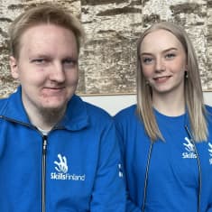 Heikki Miinalainen ja Minna Ala-Penttilä istuvat ja poseeraavat hymyillen siniset edustusasut päällä vihreällä sohvalla. Asujen rinnassa lukee SkillsFinland ja hihoissa on Suomen liput. 