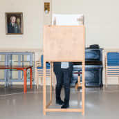 Kaksi puista äänestyskoppia suuressa tilassa, toisessa äänestyskopissa on henkilö, takaseinällä tuoleja siisteissä pinoissa, musta piano, sekä muotokuva vanhasta miehestä.