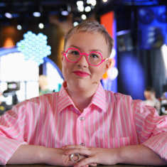 Lyhyttukkainen nainen pyöreissä pinkeissä silmälaseissa ja vaaleanpunaisessa kauluspaidassa puolikuvassa katsoo kameraan taustalla värikäs tv-studio.